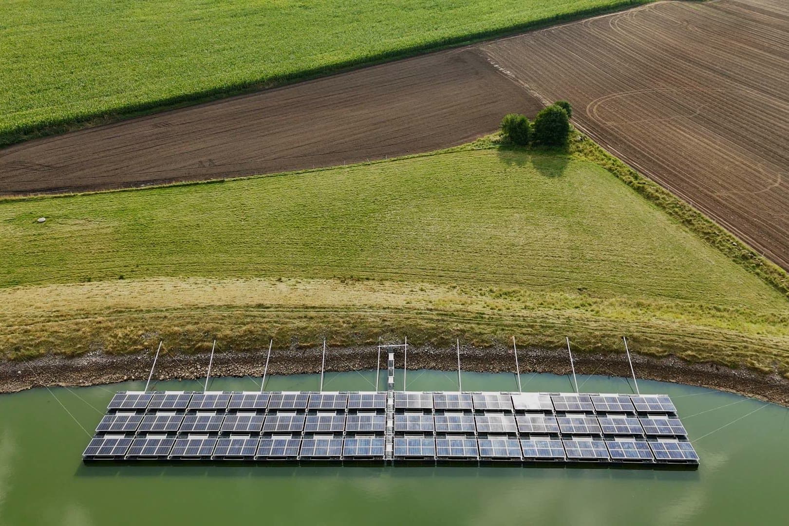 Flytande solpark längs strandkanten av en damm mellan åkrar, sett från luften